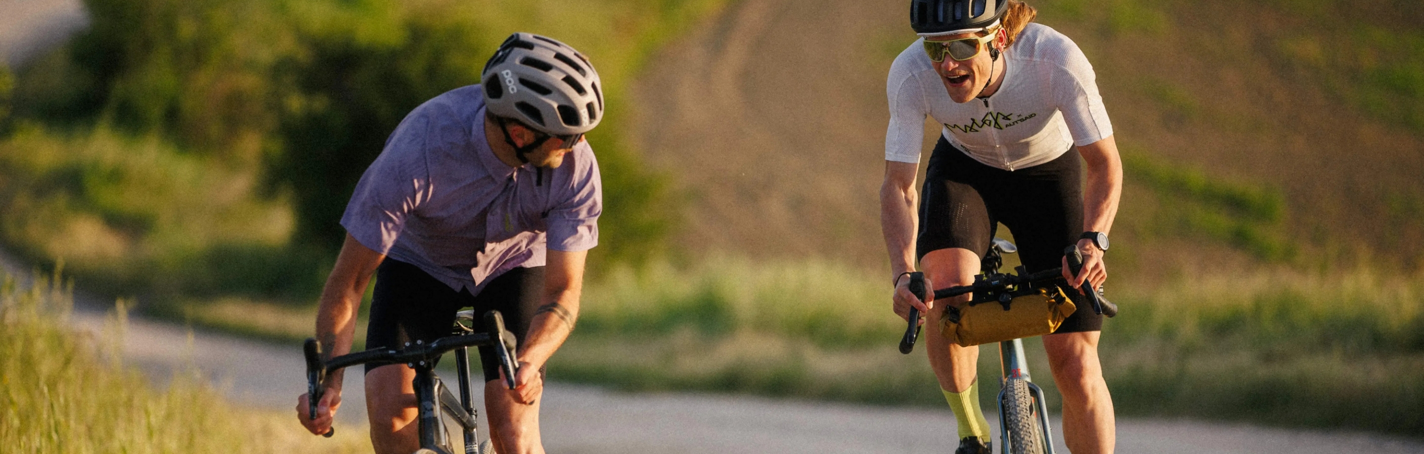 Dwóch rowerzystów gravel w letnich koszulkach rowerowych Maloja jedzie jasną ścieżką przyrodniczą w świetle wieczornym.