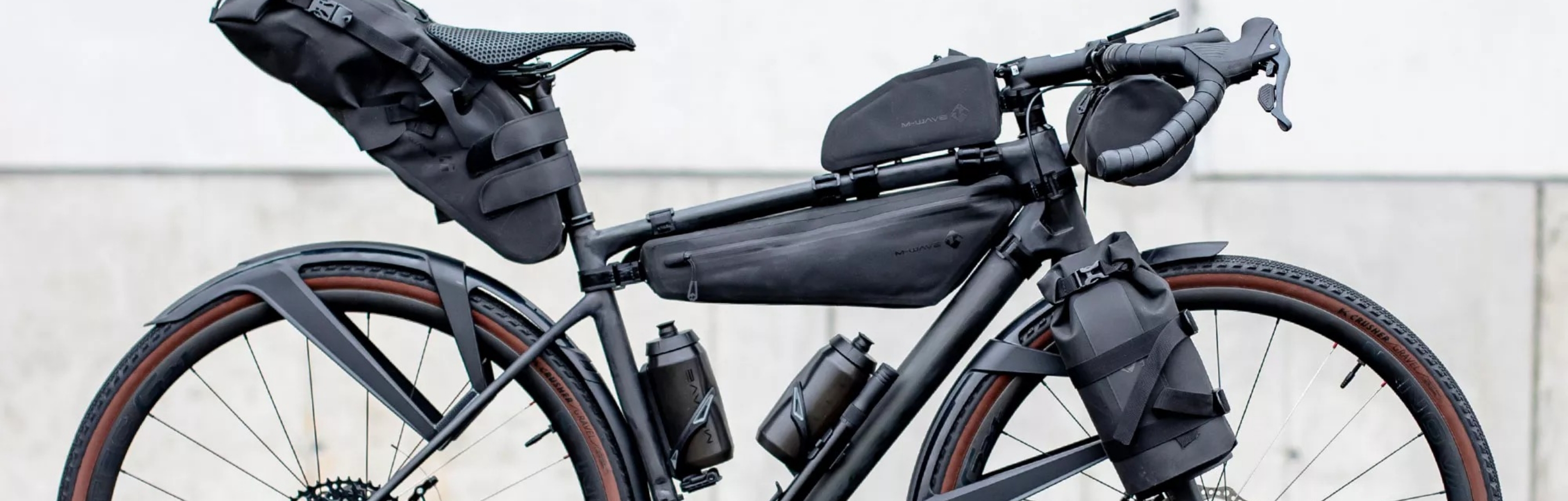 Bicicleta gravel equipada profesionalmente con una completa configuración de mochila M-Wave: bolsa de sillín, bolsa de cuadro y bolsa de manillar en negro mate para aventuras de larga distancia.