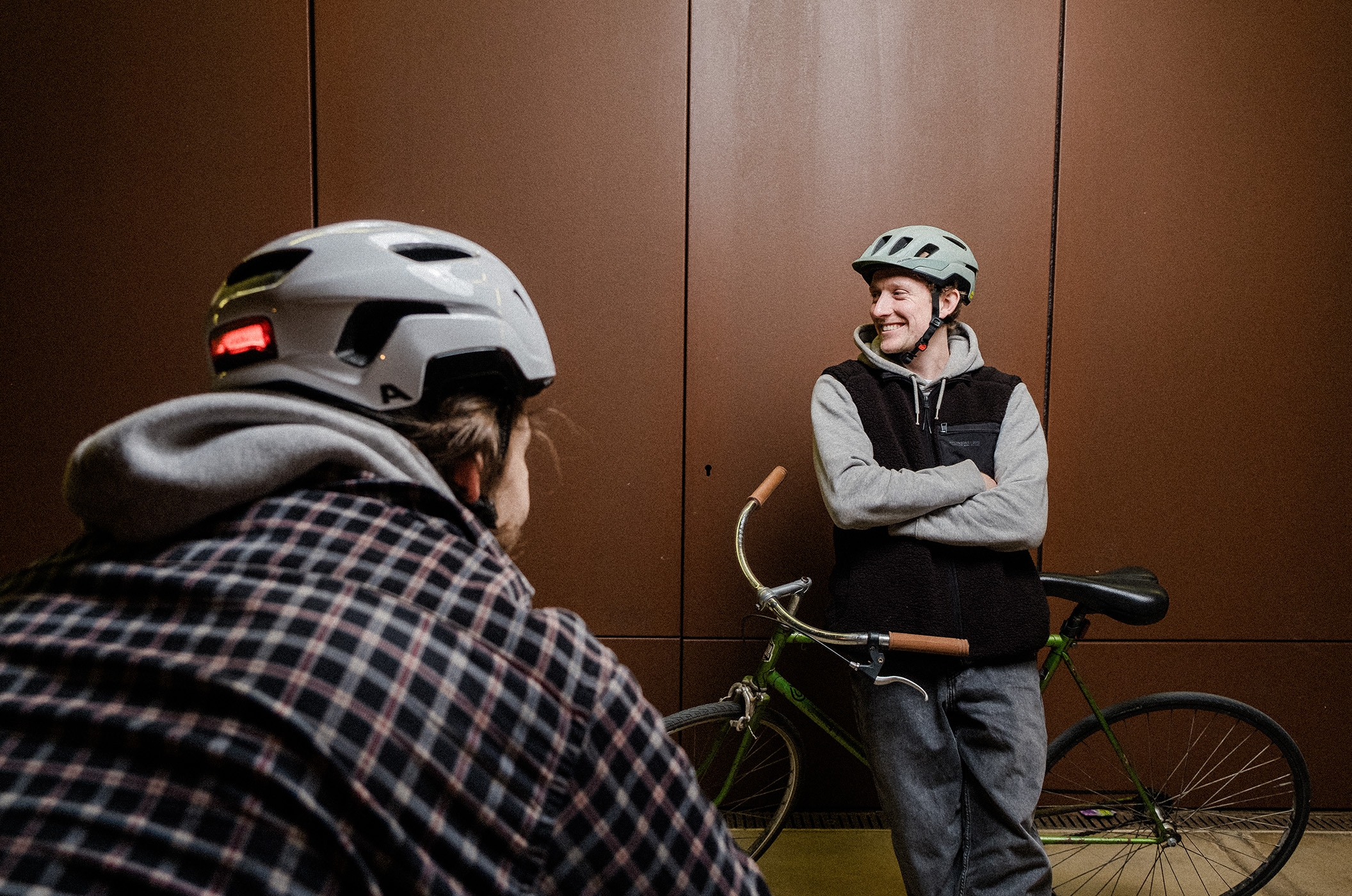 Alpina - Casques de vélo et lunettes de sport haut de gamme Deux cyclistes portant des casques vélo Alpina discutent tranquillement devant un mur marron.