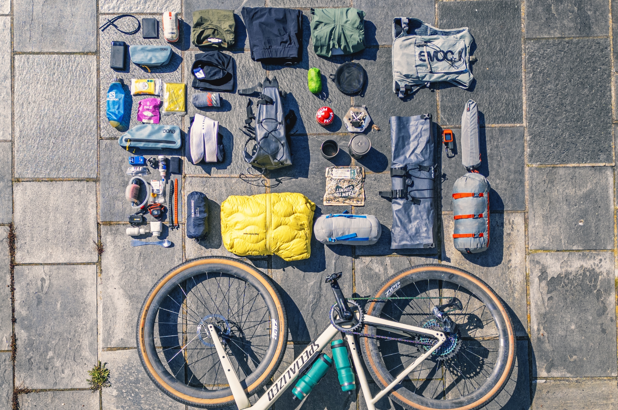Eine komplette Bikepacking-Ausrüstung – Philipp Markgraf von pedalskillcancer.com zeigt worauf es ankommt! Draufsicht auf eine vollständige Bikepacking-Ausrüstung, die am Boden ausgebreitet ist und alles von Satteltaschen, Rahmentaschen und Lenkertaschen über Schlafsysteme und Kochsets bis hin zu Werkzeugen und Trinklösungen für mehrtägige Fahrradtouren zeigt.