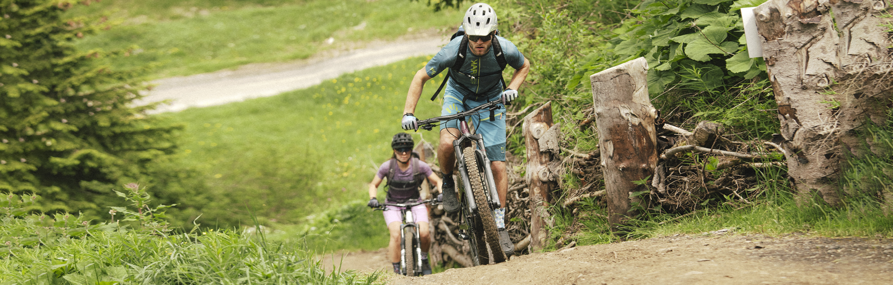 Dwóch rowerzystów górskich w odzieży rowerowej Karpos wspina się na stromą leśną ścieżkę.