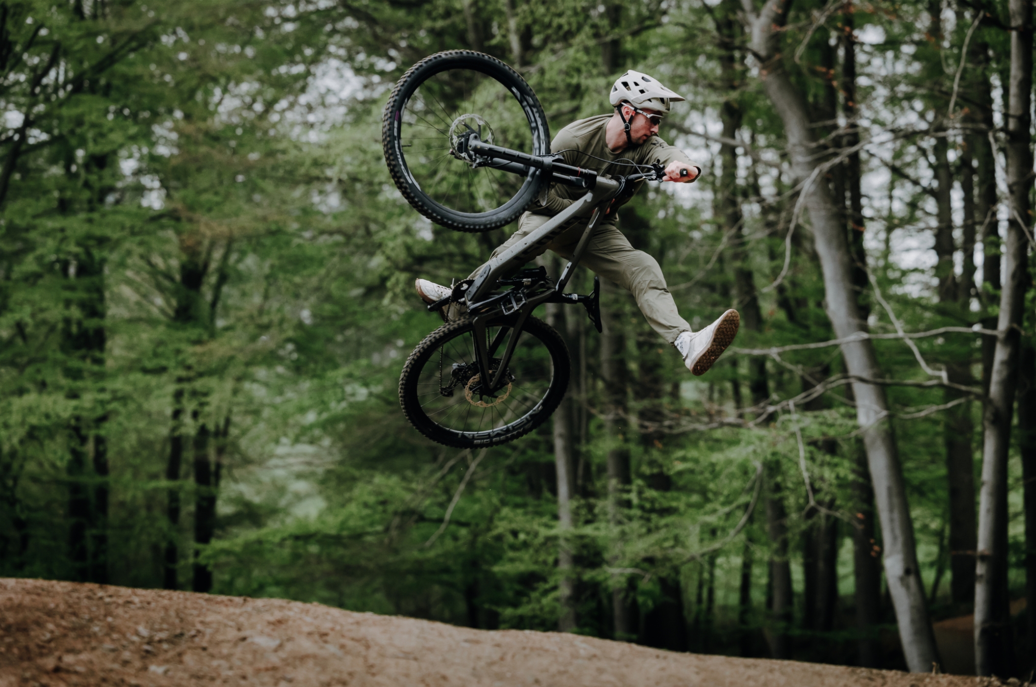 Mountainbiker auf schwarzem Ghost Bike führt einen spektakulären Whip-Jump im Wald aus, das Hinterrad zur Seite gedreht, in olivgrüner Kleidung und weißem Helm.