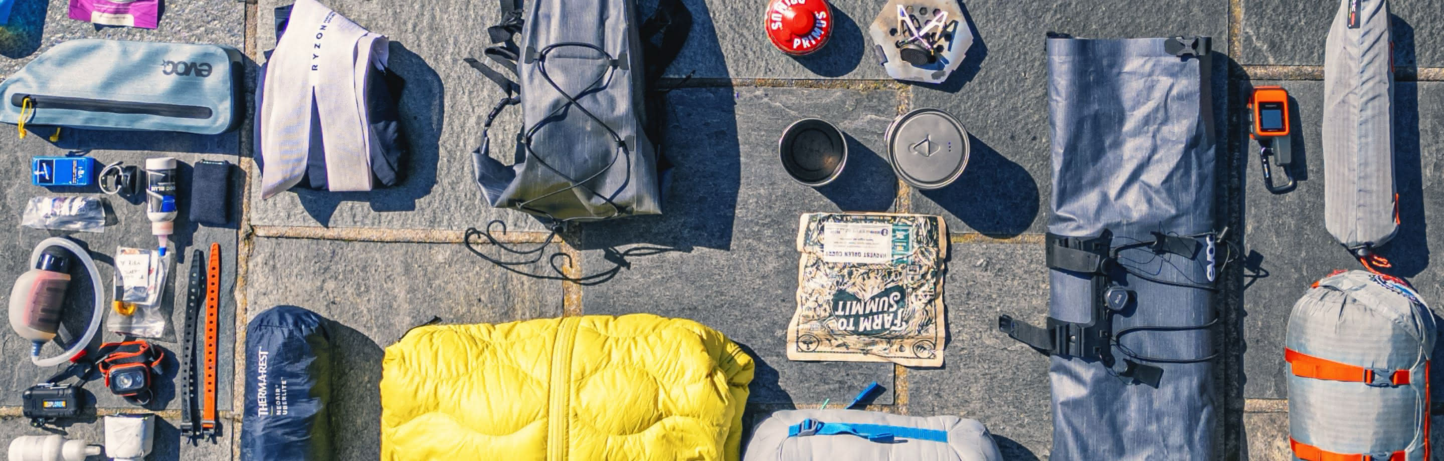 Eine sorgfältig angeordnete Auswahl an leichtem Bikepacking-Equipment für mehrtägige Radtouren, darunter ein Schlafsack, Kochutensilien, Wasserfiltersystem und kompakte Aufbewahrungslösungen auf einer Steinoberfläche.
