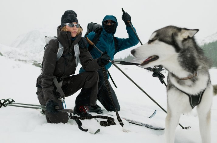 Twee wintersporters in Odlo-jassen knielen naast hun ski’s, met een husky op de voorgrond.