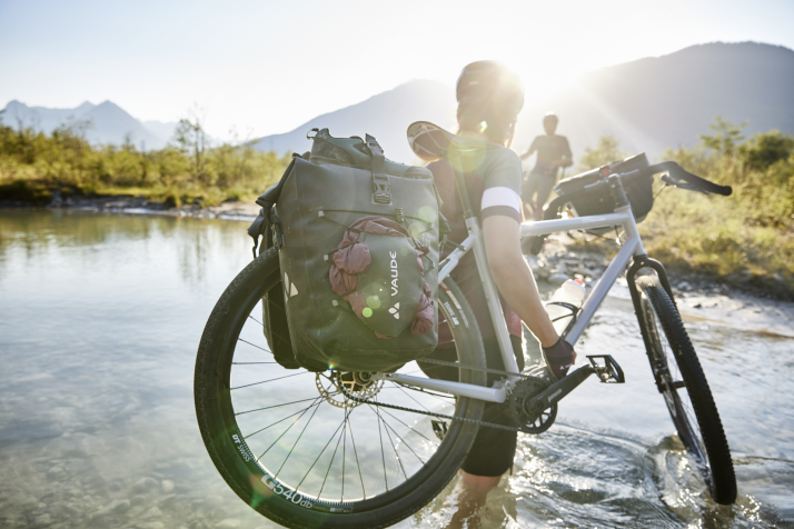 VAUDEn ”ReCycle” -pyöräilylaukut Retkipyöräilijä kantaa VAUDEn sivulaukuilla varustettua pyörää matalan joen yli aurinkoisessa vuoristomaisemassa.