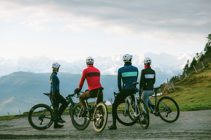 Ciclistas con Etxeondo ante montañas nevadas Cuatro ciclistas de espaldas con chaquetas de invierno Etxeondo y bicicletas de carretera frente a montañas nevadas