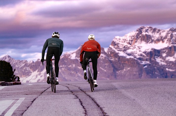 Zwei Radfahrer in Rapha Bekleidung fahren bei Sonnenuntergang eine alpine Passstraße hinauf.