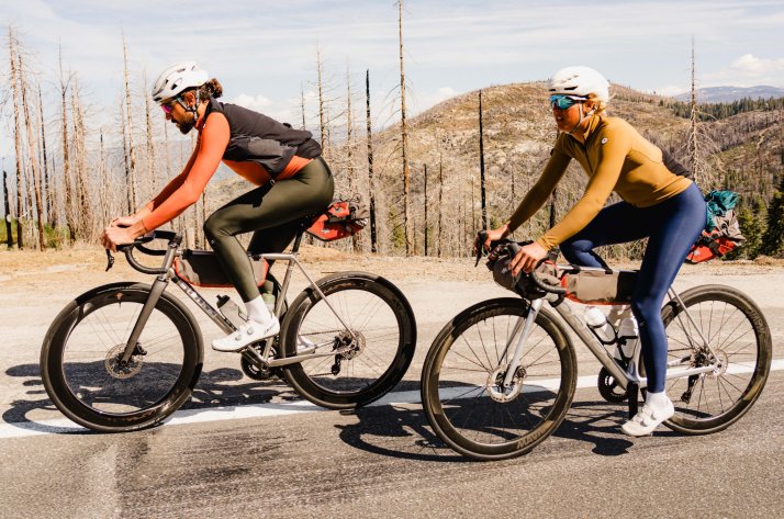 Sportlicher Fahrer in ASSOS Radtrikot und Hose, unterwegs auf regennasser Straße vor Naturkulisse.