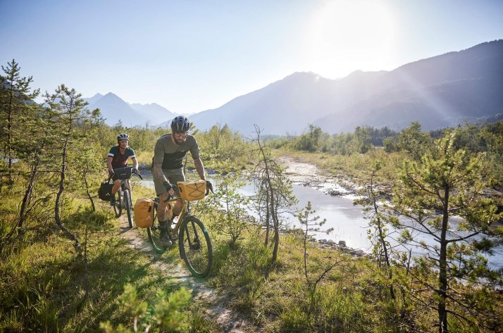 Two cyclists in VAUDE apparel riding on a narrow trail next to a river.