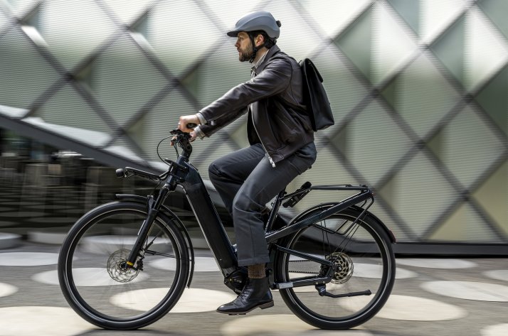 Ein Mann in urbaner Kleidung fährt auf einem schwarzen E-Bike durch eine futuristische städtische Umgebung. Er trägt eine schwarze Lederjacke, dunkle Hose und einen KASK Helm