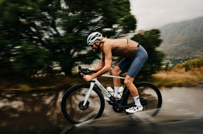 Cycliste sportif en maillot et cuissard ASSOS, roulant sur une route mouillée par la pluie dans un décor naturel.