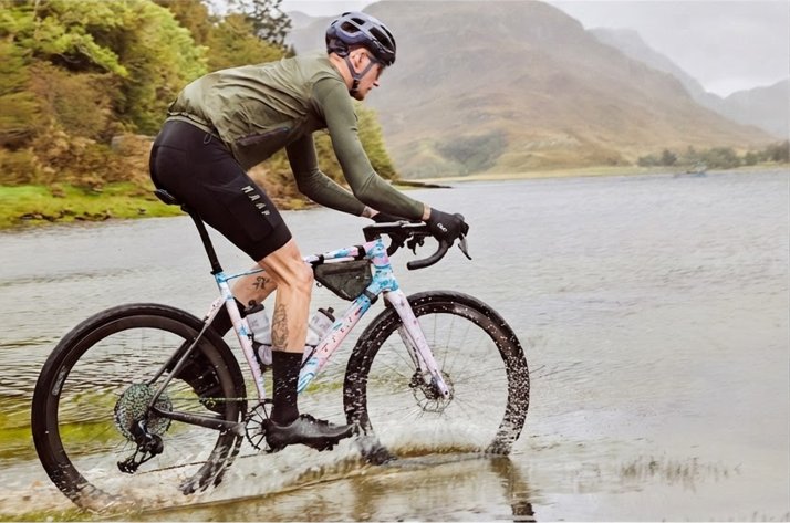  Un cycliste vêtu d’une veste MAAP traverse une rivière avec son vélo de gravel, avec les montagnes en toile de fond