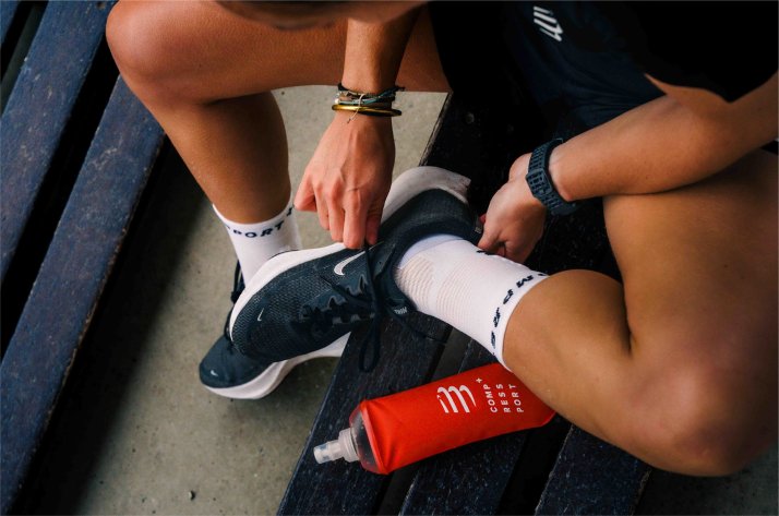Compressport-Socken und -Laufbekleidung für mehr Power Athlet schnürt Laufschuh, trägt Compressport-Kompressionssocken; im Vordergrund eine rote Compressport-Trinkflasche
