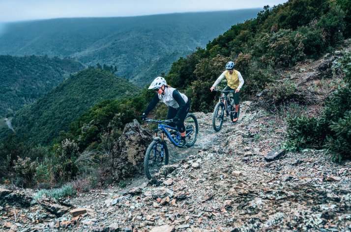 Frau und Mann in Dirtlej MTB Bekleidung fahren über einen steinigen Gebirgstrail.