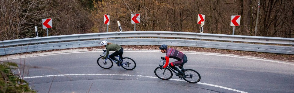 Kolarze szosowi w odzieży Rogelli pokonują zakręt na szosie przy skraju lasu.