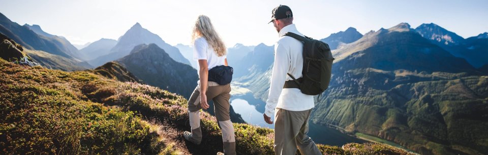 Chico y chica en la montaña haciendo senderismo con ropa Norrøna y de fondo un la lago entre montañas, día soleado