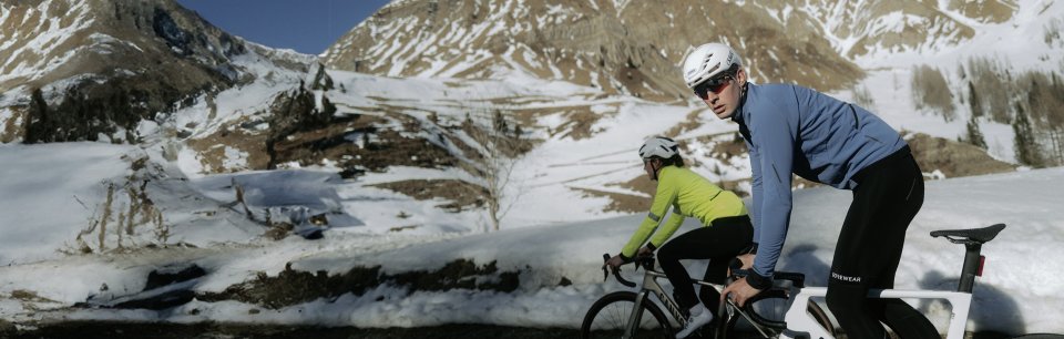 Dos ciclistas con cascos blancos pedaleando en carretera de montaña nevada, uno con maillot azul y otro con maillot verde