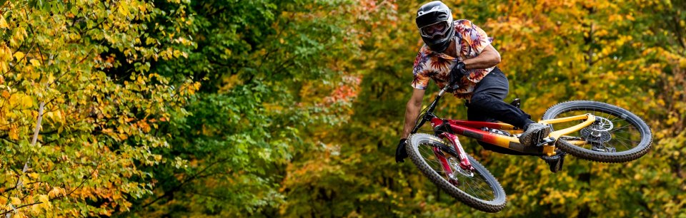 Un ciclista con una colorida camiseta de Dharco salta con su bicicleta a través de un bosque otoñal.