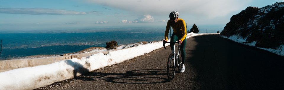 Ciclista con equipación Sportful pedaleando en carretera de montaña junto al mar, con sol y nieve en los laterales del camino