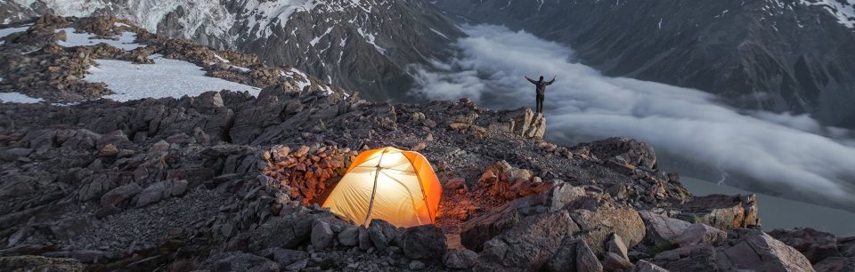 Imagen en lo alto de una montaña nevada una persona mirando al horizonte y una tienda de campaña con una linterna naranja
