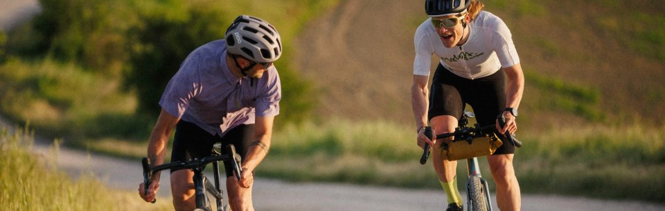 Zwei Gravel-Biker in Maloja Sommer-Radtrikots fahren auf einem hellen Naturweg bei Abendlicht.