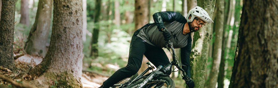 Mountain biker in iXS jersey and gloves maneuvers his bike closely past trees in the forest