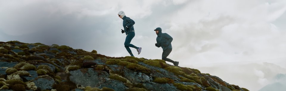 Twee trailrunners in Odlo-jassen lopen over een met mos begroeide rotskam onder een bewolkte hemel.