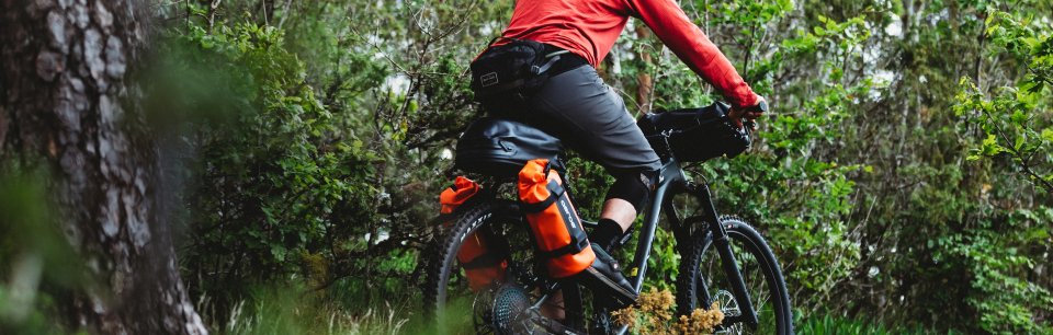 Vététiste roule avec des sacoches Aeroe orange fixées à l'arrière à travers une forêt dense