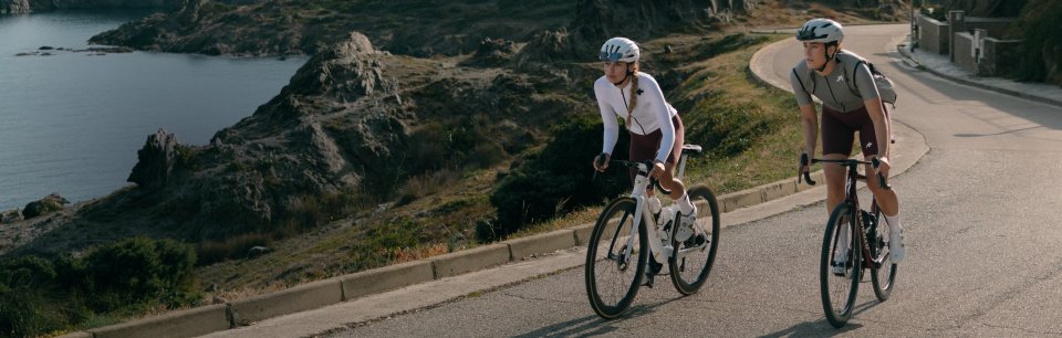 Frau und Mann in ASSOS-Outfits fahren eine Küstenstraße mit ihren Rennrädern lang