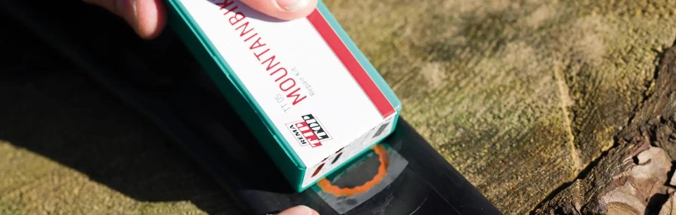 Lähikuva REMA TIP TOP -paikkaussarjan laatikosta, jota henkilö käyttää asentaessaan paikkaa polkupyörän sisäkumiin kannon päällä.