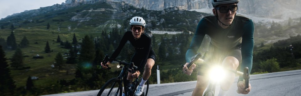 Zwei Rennradfahrende mit Lupine-Beleuchtung fahren bei Dämmerung durch eine alpine Landschaft.