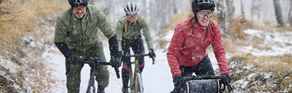 Drei Radfahrende in VAUDE Winterbekleidung fahren bei Schneefall auf einem Trail.