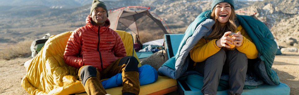 Hombre y mujer sonriendo junto a tiendas de campaña en las montañas con ropa y equipo de Mountain Hardwear.