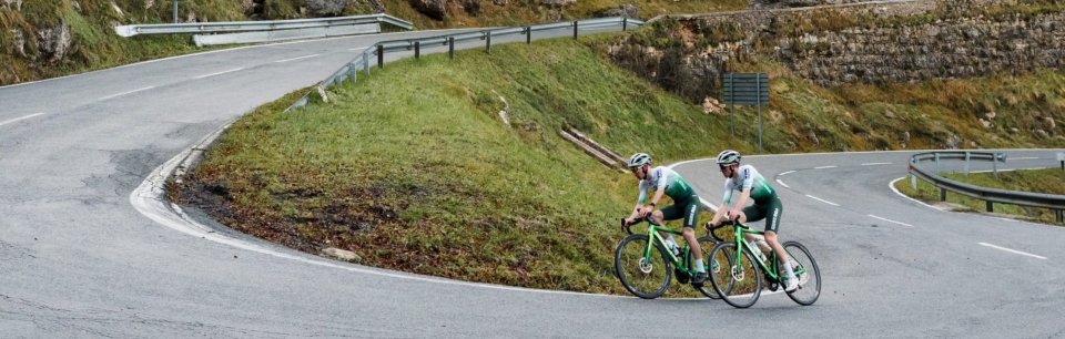 Dos ciclistas con maillots verdes pedaleando en una carretera de montaña con curvas cerradas