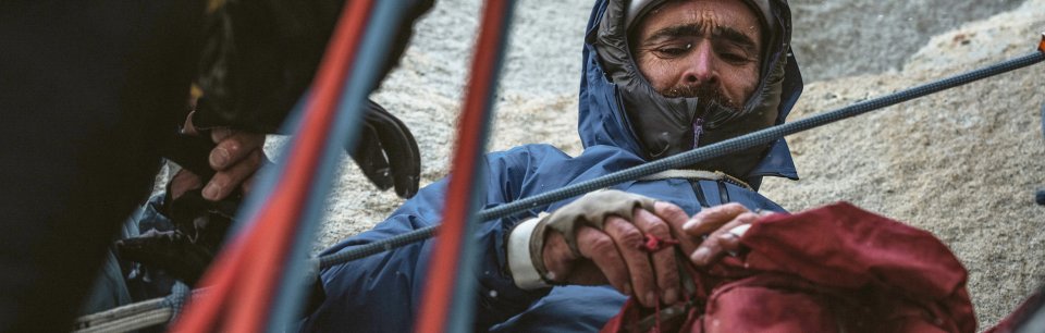 Een klimmer in een blauwe Patagonia-donsjas neemt een korte pauze, omringd door klimtouwen.