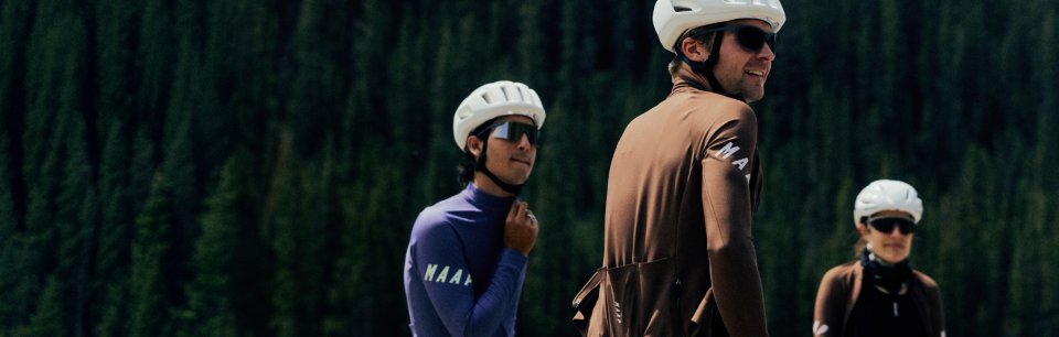 Tres ciclistas con maillots de manga larga de MAAP posan riendo uno al lado del otro frente a unos abetos frondosos