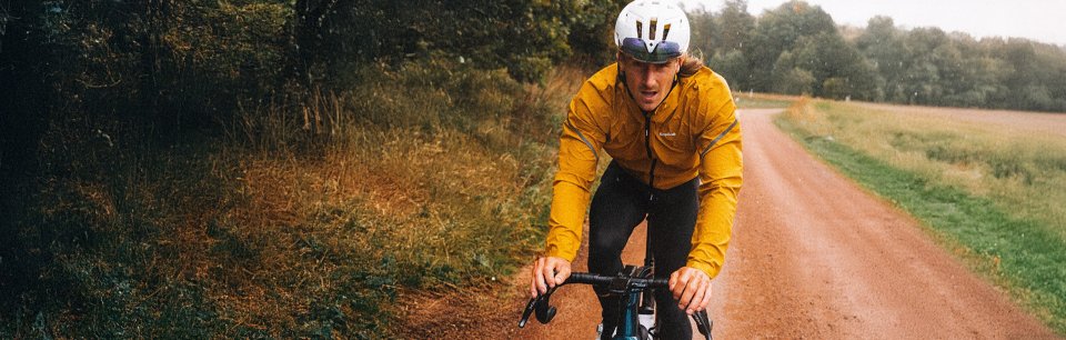 Ciclista con chaqueta amarilla y casco blanco pedaleando por un camino de gravel en un entorno natural.