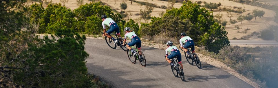 Vier Rennradfahrer in Milremo Radbekleidung fahren in einer Gruppe eine kurvige Landstraße entlang